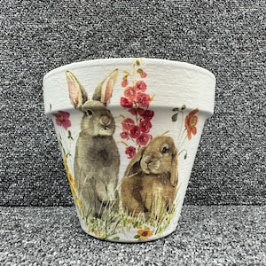 May include: White ceramic flower pot featuring a watercolor design of two rabbits, one upright and one seated, amidst wildflowers and red floral accents. The pot has a textured finish, suitable for indoor or outdoor use.