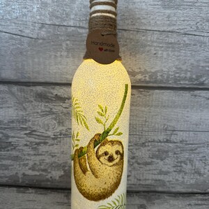 May include: A white glass bottle with a sloth design and green leaves. The bottle is decorated with twine and a brown tag that says "Handmade with Love".
