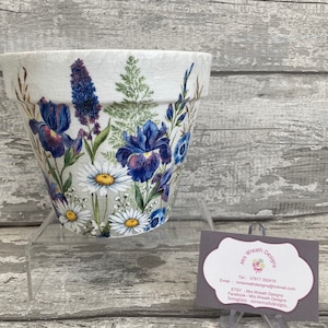 May include: A white ceramic plant pot featuring a floral design with blue irises and daisies. The pot is displayed on a clear stand. A business card with the text "Mrs Wreath Designs" is also visible.