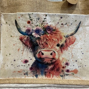 K&ouml;nnte beinhalten: Eine braune und wei&szlig;e Highland-Kuh mit einer Blumenkrone auf dem Kopf. Die Kuh ist in Aquarelltechnik mit Farbspritzern auf wei&szlig;em Hintergrund gemalt. Das Bild befindet sich auf einer Jute-Tragetasche.