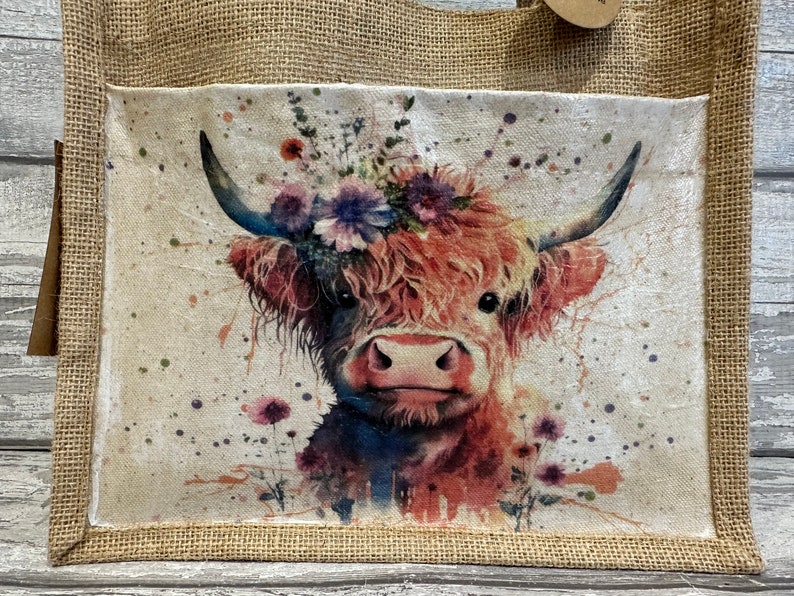 K&ouml;nnte beinhalten: Eine braune und wei&szlig;e Highland-Kuh mit einer Blumenkrone auf dem Kopf. Die Kuh ist in Aquarelltechnik mit Farbspritzern auf wei&szlig;em Hintergrund gemalt. Das Bild befindet sich auf einer Jute-Tragetasche.
