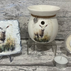 May include: A white ceramic wax melt warmer featuring a lamb design. The warmer is on a glass stand and has a small heart charm hanging from it. There is a white wax melt in a clear container and a white tea light candle on a silver holder.