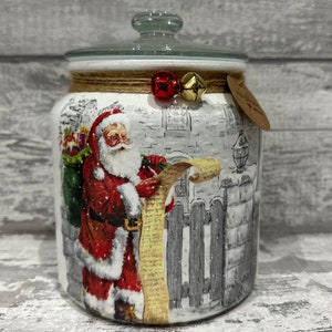 Peut inclure: Pot en céramique blanche avec un couvercle en verre et une scène de Noël représentant le Père Noël tenant une liste. Le pot est décoré d'un nœud en ficelle brune avec des clochettes rouges et dorées.