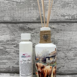 Peut inclure: Un flacon diffuseur blanc avec une scène de paysage de vaches des Highlands, une maison et des fleurs. Le flacon a un col en jute, un charme en forme de cœur et des roseaux en bois. Une bouteille blanche de recharge de diffuseur "Fresh Linen" est à côté.