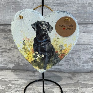 May include: A black Labrador Retriever dog is pictured on a heart-shaped slate plaque. The plaque is on a black metal stand. The plaque has a brown tag that says "Handmade with love".