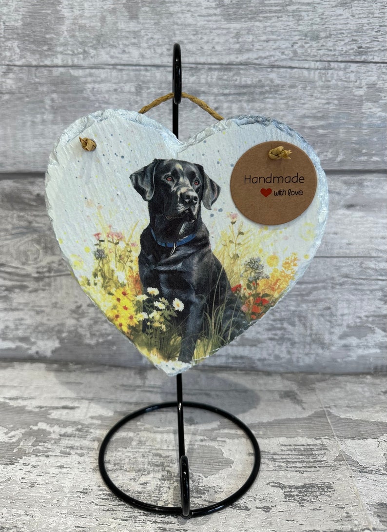 May include: A black Labrador Retriever dog is pictured on a heart-shaped slate plaque. The plaque is on a black metal stand. The plaque has a brown tag that says "Handmade with love".