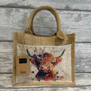 K&ouml;nnte beinhalten: Eine braune Jute-Tragetasche mit einem Blumendesign, das eine Highland-Kuh zeigt. Die Tasche hat eine Tasche auf der Vorderseite und ein Etikett mit der Aufschrift "Westford Mill Jute".