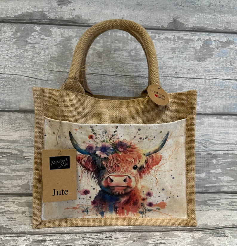 K&ouml;nnte beinhalten: Eine braune Jute-Tragetasche mit einem Blumendesign, das eine Highland-Kuh zeigt. Die Tasche hat eine Tasche auf der Vorderseite und ein Etikett mit der Aufschrift "Westford Mill Jute".