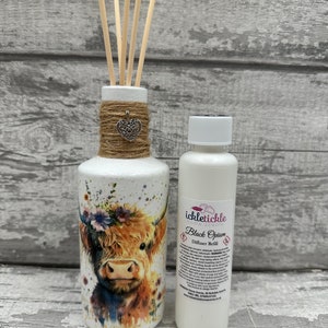 May include: White ceramic diffuser bottle featuring a Highland Cow design, adorned with a brown twine bow and a silver heart charm. The bottle is filled with wooden diffuser sticks. Beside it is a white bottle with a black label reading "ickletickle home scents Black Opium Diffuser Refill".