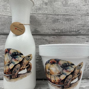 Könnte beinhalten: Zwei weiße Keramik-Blumentöpfe mit einem gemalten Bild einer Schildkröte. Der größere Topf ist eine Vase mit einem braunen Etikett, auf dem "Handgefertigt mit Liebe" steht.