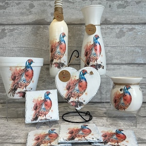 May include: A set of home decor items featuring a watercolor design of a pheasant. The set includes a vase, a bottle, a planter, a heart-shaped slate, a candle warmer, and four coasters. The items are all white with a watercolor design of a pheasant in shades of blue, brown, and red.