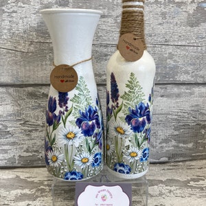 May include: Two white ceramic vases with floral designs. The vases are decorated with blue and white irises, daisies, and other flowers. The vases are handmade and have a rustic charm. The text "Handmade with love" is on a tag attached to each vase.