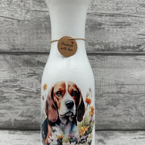 May include: White ceramic vase featuring a beagle dog and floral design. The vase has a brown tag that reads "Handmade with love".