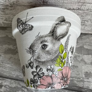 May include: White ceramic planter featuring a hand-painted design of a rabbit, flowers, and a butterfly. The rabbit is grey and white, the flowers are pink and green, and the butterfly is black and white.