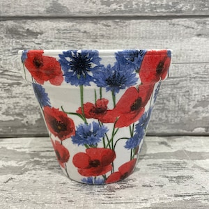 May include: A white ceramic flower pot with a red and blue floral pattern. The pot is decorated with red poppies and blue cornflowers.