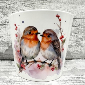 May include: White ceramic planter featuring a watercolor illustration of two robins perched on a branch with red berries. The birds have orange breasts, brown wings, and black beaks. The planter has a slightly flared shape.