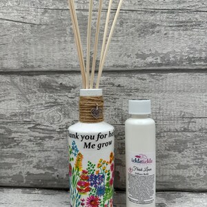 Peut inclure: Bouteille de diffuseur en céramique blanche avec un motif floral et le texte "Merci de m'aider à grandir". La bouteille est remplie de bâtonnets diffuseurs en bois et est ornée d'un nœud en ficelle brune. Une bouteille de recharge de diffuseur blanche est placée à côté de la bouteille de diffuseur. La bouteille de recharge est dotée d'une étiquette indiquant "ickletickle home scents" et "Recharge de diffuseur Linge frais".