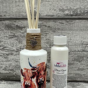 Peut inclure: Une bouteille de diffuseur blanche avec un nœud en ficelle brune et un charme de cœur argenté. La bouteille présente une peinture à l'aquarelle de deux vaches des Highlands. Une petite bouteille blanche avec une étiquette noire qui indique "ickletiickle Home Scents Black Opium Diffuser Refill" est à côté de la bouteille de diffuseur.