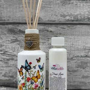May include: A white diffuser bottle with a butterfly and floral design, wrapped with twine and a heart charm. The bottle holds wooden reeds. A white bottle of "Fresh Linen" diffuser refill is next to it.