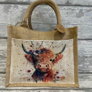 K&ouml;nnte beinhalten: Eine braune Jute-Tragetasche mit einer wei&szlig;en Stofftasche, die ein Aquarellbild einer Highland-Kuh mit einer Blumenkrone zeigt. Die Tasche hat ein braunes Papieretikett mit der Aufschrift "Handmade with love".