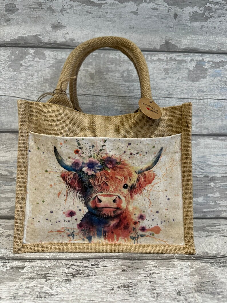 K&ouml;nnte beinhalten: Eine braune Jute-Tragetasche mit einer wei&szlig;en Stofftasche, die ein Aquarellbild einer Highland-Kuh mit einer Blumenkrone zeigt. Die Tasche hat ein braunes Papieretikett mit der Aufschrift "Handmade with love".
