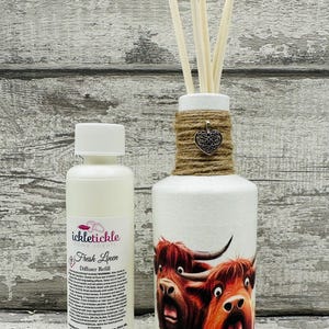 Peut inclure: Une bouteille de diffuseur blanche avec un enroulement de ficelle brune et un charme de cœur argenté. La bouteille présente une image de dessin animé de deux vaches des Highlands avec des expressions surprises. Une petite bouteille blanche avec une étiquette qui indique "ickletiickle Home Scents Fresh Linen Diffuser Refill" est à côté de la bouteille de diffuseur.