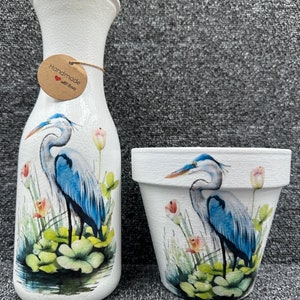 May include: Two white ceramic vases featuring a blue heron design. The vase on the left is taller and has a tag that reads "Handmade with love". The vase on the right is shorter and wider. Both vases have a watercolour-style design of a blue heron standing in a marsh.