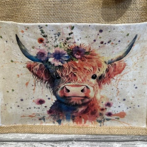 K&ouml;nnte beinhalten: Eine braune und wei&szlig;e Highland-Kuh mit einer Blumenkrone, im Aquarell-Stil gemalt, auf einem wei&szlig;en Leinwandhintergrund mit braunen Spritzern. Das Bild befindet sich auf einer Jute-Tragetasche.