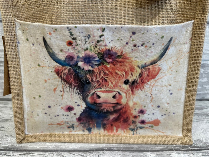 K&ouml;nnte beinhalten: Eine braune und wei&szlig;e Highland-Kuh mit einer Blumenkrone, im Aquarell-Stil gemalt, auf einem wei&szlig;en Leinwandhintergrund mit braunen Spritzern. Das Bild befindet sich auf einer Jute-Tragetasche.