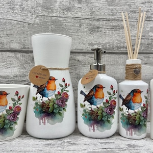 May include: A white ceramic bathroom set with a robin bird and floral design. The set includes a tumbler, vase, soap dispenser, diffuser, and a small bottle. Each item features a watercolour-style illustration.