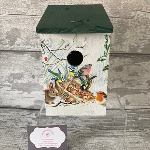 May include: A wooden birdhouse with a green roof and a white base. The birdhouse is decorated with a winter scene featuring birds, a rabbit, and berries. The birdhouse has a round hole for birds to enter. The birdhouse is sitting on a clear acrylic stand. The image also includes a business card for "Mrs Wreath Designs".