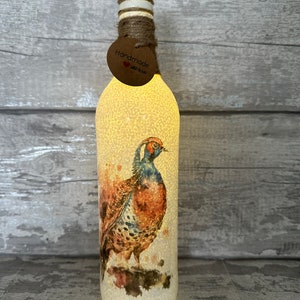 May include: A white glass bottle with a brown pheasant design and a cork top. The bottle is lit from within, creating a warm glow. The bottle has a handmade tag with the words "Handmade with Love".