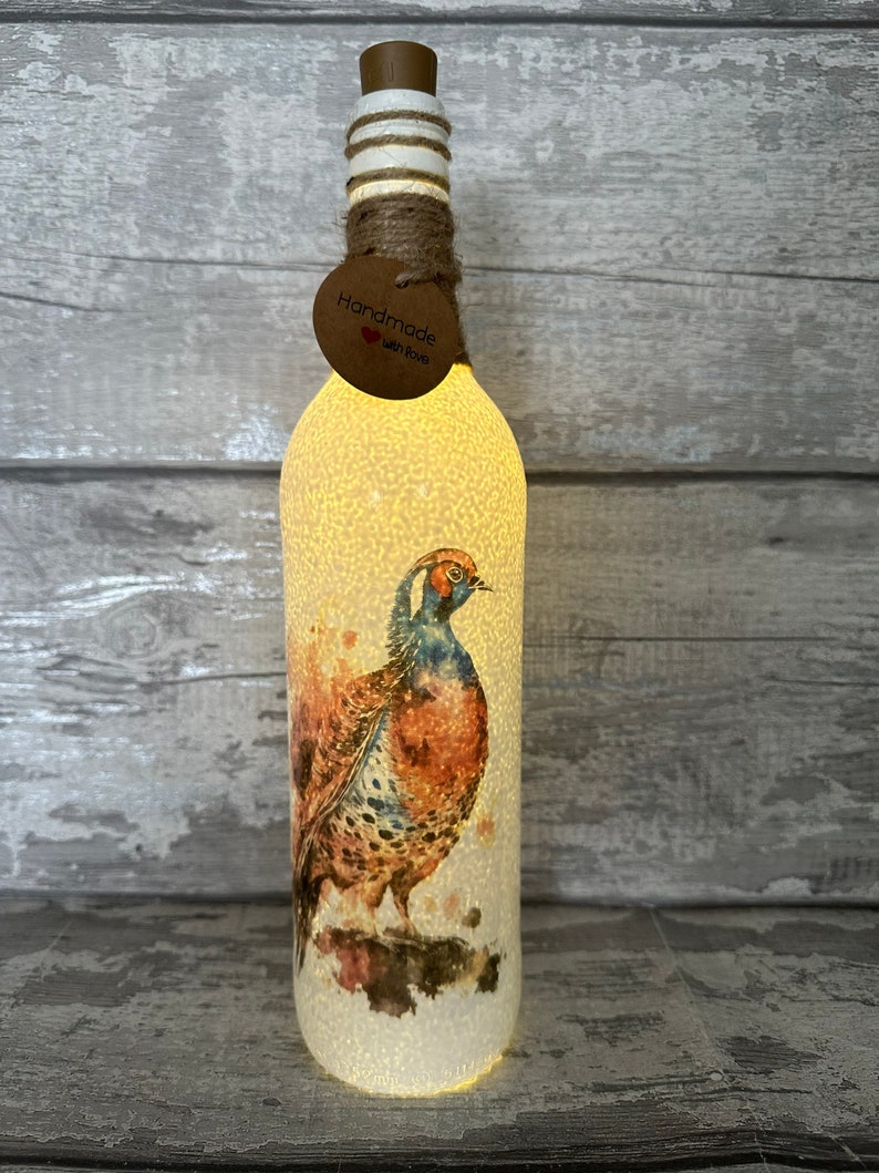 May include: A white glass bottle with a brown pheasant design and a cork top. The bottle is lit from within, creating a warm glow. The bottle has a handmade tag with the words "Handmade with Love".