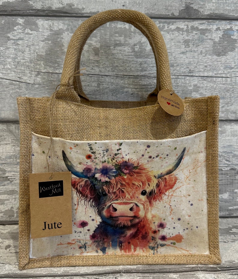 K&ouml;nnte beinhalten: Eine braune Jute-Tragetasche mit einem Blumenmuster einer Highland-Kuh. Die Tasche hat eine Tasche auf der Vorderseite mit einem Etikett, auf dem "Westford Mill Jute" steht. Die Tasche hat auch ein Etikett mit der Aufschrift "Handmade with love".
