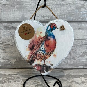 May include: Handmade with love heart shaped slate ornament featuring a watercolor painting of a pheasant. The ornament is on a black metal stand.