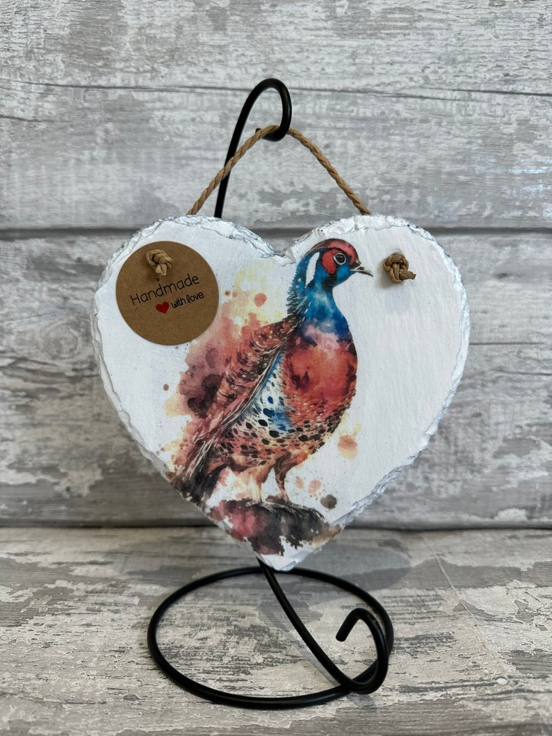 May include: Handmade with love heart shaped slate ornament featuring a watercolor painting of a pheasant. The ornament is on a black metal stand.