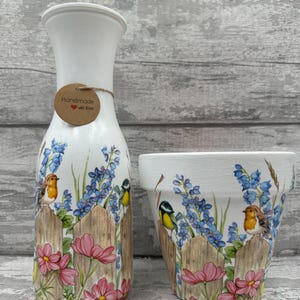 Peut inclure: Un vase en céramique blanc et un pot assorti décorés d'une scène champêtre. Le motif comprend une clôture en bois, des fleurs roses, des fleurs sauvages bleues et des oiseaux colorés. Le vase porte une étiquette "Handmade with love".
