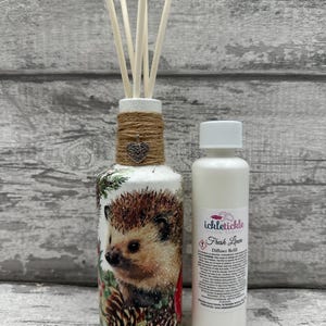 May include: A white glass bottle with a brown twine bow and a silver heart charm. The bottle is decorated with a hedgehog and pine cones. The bottle is filled with diffuser sticks. A white bottle with a white label that reads "ickletickle home scents Fresh Linen Diffuser Refill" is next to the bottle.