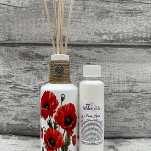 Peut inclure: Un flacon diffuseur en céramique blanche avec un motif floral de coquelicots rouges, des bâtonnets en bois et une petite bouteille blanche étiquetée "Fresh Linen Diffuser Refill". Le flacon est entouré de ficelle et d'un charm en forme de cœur.