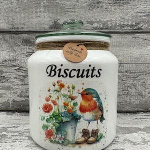 May include: White ceramic biscuit jar with a green glass lid. The jar features a robin bird illustration with a bucket of flowers and the text "Biscuits" in black lettering. A brown tag with the text "Handmade with love" is attached to the jar.