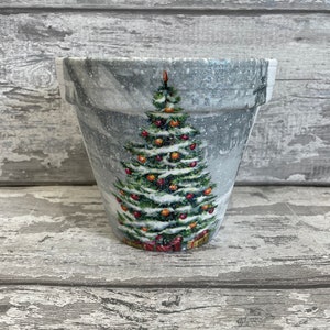 May include: A white ceramic planter with a winter scene featuring a green Christmas tree with red baubles and presents. The planter has a snowy, textured surface.