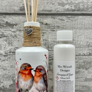 May include: White ceramic reed diffuser featuring a watercolour painting of two robins. The neck is wrapped in jute, with a silver heart charm. A white bottle of diffuser refill is next to it.