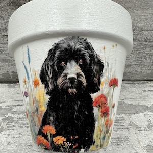 May include: White painted flower pot with a black dog portrait, set against a backdrop of wildflowers in various colours. The dog's image is the focal point. A decorative item for home or garden, suitable for planting.