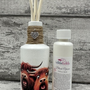 Peut inclure: Un diffuseur d'huiles essentielles en céramique blanche avec un charme en forme de cœur décoratif et des bâtonnets en bois. Le diffuseur présente un motif de vache des Highlands. Une bouteille assortie de recharge de diffuseur "Black Opium" est à côté.