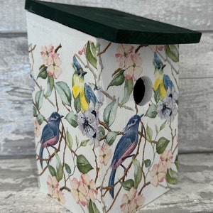 May include: A white wooden birdhouse with a green roof. The birdhouse is decorated with a floral pattern featuring pink and white flowers, green leaves, and blue birds.