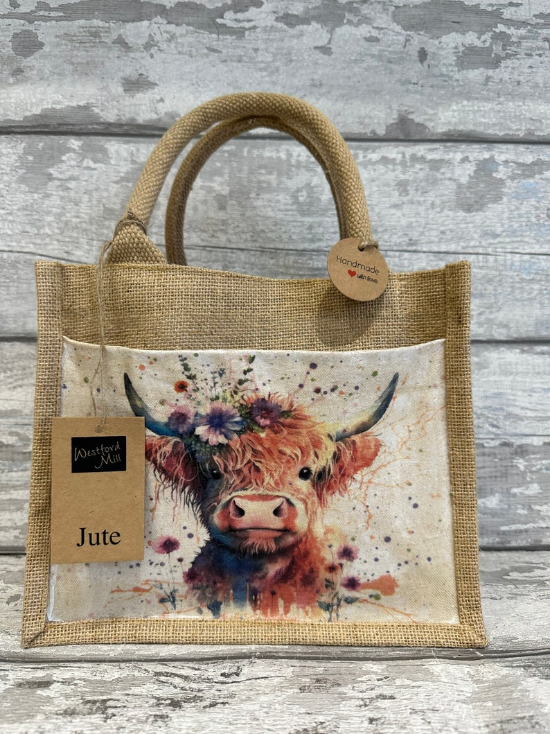 K&ouml;nnte beinhalten: Eine braune Jute-Tragetasche mit einem Blumendesign, das eine Highland-Kuh zeigt. Die Tasche hat ein braunes Papieretikett mit der Aufschrift "Westford Mill" und "Jute". Ein kleines braunes Papieretikett in Kreisform lautet "Handmade with love".