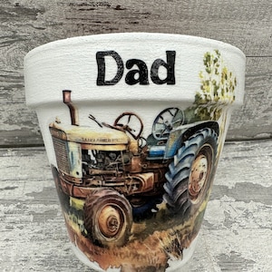 May include: White ceramic planter with a blue and yellow tractor image and the word "Dad" written in black lettering.