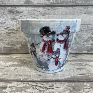 Könnte beinhalten: Ein weißer Keramiktopf mit einer Winterszene mit drei Schneemännern. Die Schneemänner tragen Hüte und Schals und sind von Schnee umgeben. Der Topf ist mit einer blauen und weißen Glasur verziert.
