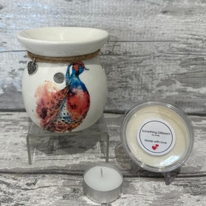 May include: White ceramic wax melt burner with a watercolor pheasant design. The burner has a small hole in the top for a tea light. The burner is on a clear stand. A small jar of wax melts is next to the burner. The jar has a white label that says "Something Different! by Linda" and "Made with love". A tea light is in front of the burner.