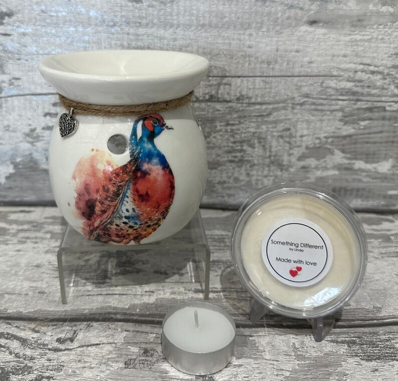 May include: White ceramic wax melt burner with a watercolor pheasant design. The burner has a small hole in the top for a tea light. The burner is on a clear stand. A small jar of wax melts is next to the burner. The jar has a white label that says "Something Different! by Linda" and "Made with love". A tea light is in front of the burner.